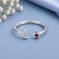 A silver open ring featuring a crescent moon half-set with small clear stones and a five-pointed star set with a round red gemstone, displayed on a light blue fabric background with dried white flowers in the corner.