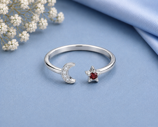 A silver open ring featuring a crescent moon half-set with small clear stones and a five-pointed star set with a round red gemstone, displayed on a light blue fabric background with dried white flowers in the corner.