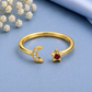 A gold open-band ring with a crescent moon charm set with small clear stones and a star charm set with a red gemstone, displayed on a blue fabric background with white flowers and a blue satin cloth.