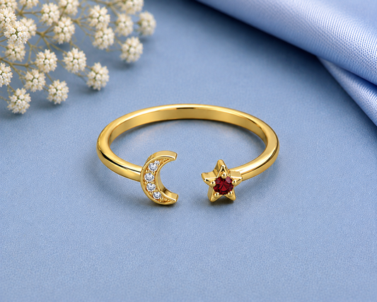 A gold open-band ring with a crescent moon charm set with small clear stones and a star charm set with a red gemstone, displayed on a blue fabric background with white flowers and a blue satin cloth.