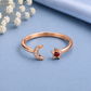 A rose gold open bangle ring featuring a crescent moon charm set with small clear stones and a five-pointed star charm set with a red gemstone, displayed on a textured light blue surface with dried white flowers in the corner.